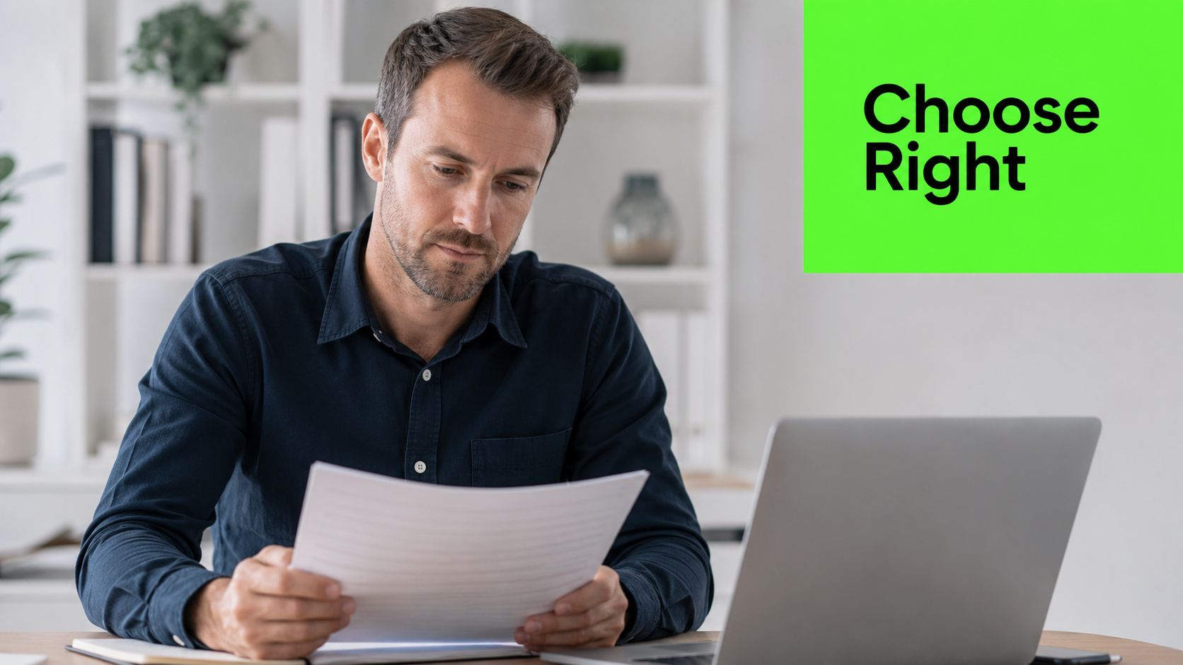 A professional man reading documents while working at a laptop in a bright office environment.